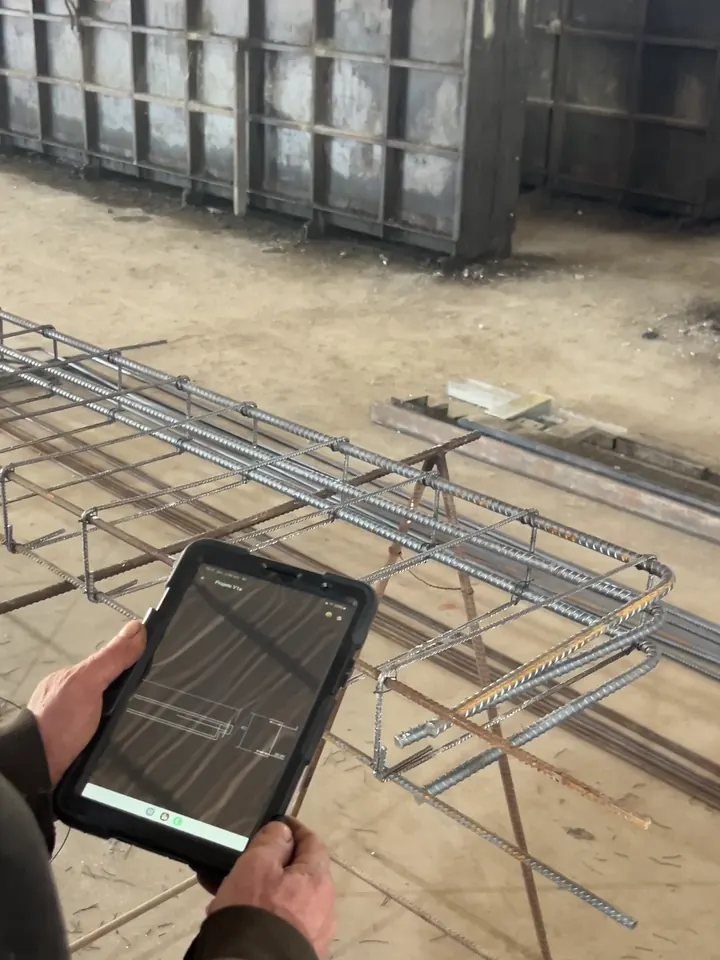 Armador da Gemold conferindo o desenho da viga no tablet ao lado da armadura montada, com as formas metálicas ao fundo